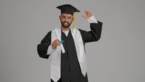 جودة التعليم في الامارات، الوصول لحلم النجاح، تعابير السعادة والسرور، مناسبة النجاح في الجامعة، الاحتفال بإنهاء الدراسة الجامعية، مفهوم الاجتهاد والتفوق، تصوير لشاب خريج عربي خليجي اماراتي يرتدي عباءة وقبعة التخرج يمسك بيده الشهادة، يميل رأسه بايماءات الفخر ، خلفية رماديه