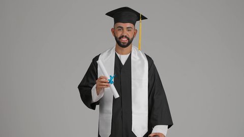 جودة التعليم في الامارات، الوصول لحلم النجاح، تعابير السعادة والسرور، مناسبة النجاح في الجامعة، الاحتفال بإنهاء الدراسة الجامعية، مفهوم الاجتهاد والتفوق، تصوير لشاب خريج عربي خليجي اماراتي مبتسم يرتدي عباءة وقبعة التخرج  يمسك بيده الشهادة، خلفية رماديه