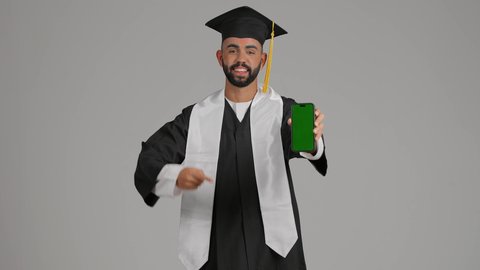 عرض شاشة خضراء كروما على الهاتف المحمول، برامج وتطبيقات التعليم والدراسة اونلاين، تصوير لشاب خريج عربي خليجي اماراتي مبتسم يرتدي القبعة وزي التخرج يمسك بيده الجوال ، الاحتفال بانتهاء المرحلة الجامعية، خلفية رماديه