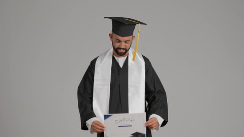 The concept of diligence and excellence, expressions of happiness and pleasure, a university occasion and celebration, achieving the dream of success, celebrating the completion of university studies, a picture of a smiling young Arab Gulf Emirati graduate wearing a graduation gown and holding a certificate in his hand, gray background
