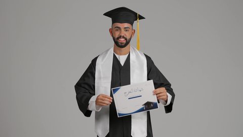 تعابير السعادة والسرور، مناسبة واحتفال جامعي، الوصول لحلم النجاح، الاحتفال بإنهاء الدراسة الجامعية، مفهوم الاجتهاد والتفوق، تصوير لشاب خريج عربي خليجي اماراتي مبتسم يرتدي عباءة التخرج يمسك بيده الشهادة، خلفية رماديه