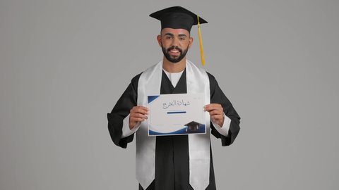 الوصول لحلم النجاح، تعابير السعادة والسرور، مناسبة واحتفال جامعي، الاحتفال بإنهاء الدراسة الجامعية، مفهوم الاجتهاد والتفوق، تصوير لشاب خريج عربي خليجي اماراتي مبتسم يرتدي عباءة التخرج يمسك بيده الشهادة، خلفية رماديه