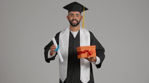 Obtaining a graduation certificate, giving gifts on occasions, celebrating the completion of university studies, gestures of happiness for reaching the dream of success, a picture of a young Arab Gulf Emirati graduate wearing a graduation outfit and cap, holding a certificate and gift box, gray background