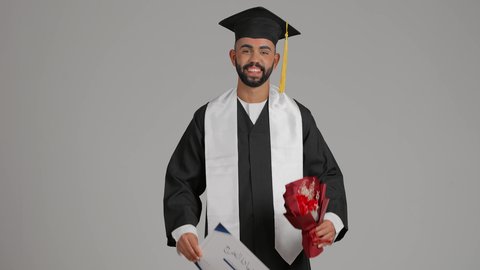 الشعور بالفخر والثقة بالنفس، استلام الشهادة بتعابير الفرح والسرور، مفهوم التفوق والاجتهاد، تصوير لخريج عربي خليجي اماراتي يرتدي قبعة وثوب التخرج يحمل باقة الورد بايماءات السعادة، الاحتفال بالنجاح وانهاء الدراسة الجامعية، خلفية رماديه