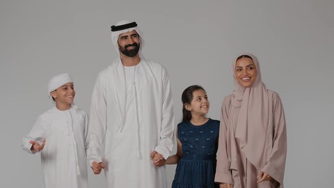 مفهوم الأمان و الاستقرار العائلي، أجواء عائلية سعيدة، الاشارة إلى شيء ما بعيد والنظر إليه بإيماءات الاعجاب، المشي و التنزه مع الأطفال، بورتريه لام عربيه خليجيه اماراتية ترتدي العباءه الملونه تقوم بقضاء وقت ممتع برفقه اولادها و زوجها ، خلفية رمادية
