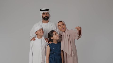 الاشارة إلى شيء ما بعيد والنظر إليه بإيماءات الاعجاب، مفهوم الأسرة الإماراتية السعيدة، المشي و التنزه مع الأطفال، مفهوم الترابط الأسري، بورتريه لام عربيه خليجيه اماراتية ترتدي العباءه الملونه تقوم بقضاء وقت ممتع برفقه اولادها و زوجها ، خلفية رمادية