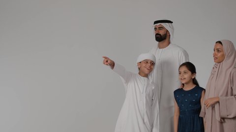 عائله عربيه خليجيه اماراتية ترتدي الزي التقليدي تقوم بالمشي و التنزه ، مفهوم الترابط الأسري، مشاركة الأوقات الممتعة مع الأبناء، بورتريه لطفل عربي خليجي  يقوم بالاشارة إلى شيء ما بعيد والنظر إليه، خلفية رمادية