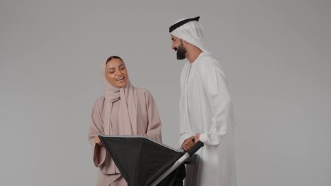القيام بالنزهات والجولات العائلية، مفهوم الأمومة والرعايه والاهتمام بالأولاد، بورتريه لام عربيه خليجيه اماراتية ترتدي العباءه الملونه برفقه زوجها تنظر الى إبنها الرضيع داخل العربة و تلعب معه بسعادة بإيماءات تدل على الحب والأمان