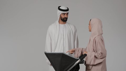 عائلة اماراتية تقضي اوقات ممتعة، الرعايه والاهتمام بالأولاد، بورتريه لأب عربي خليجي اماراتي يرتدي الكندورة و الغتره برفقه زوجته ينظر الى إبنه الرضيع داخل العربة و يلعب معه بسعادة بإيماءات تدل على الحب والأمان