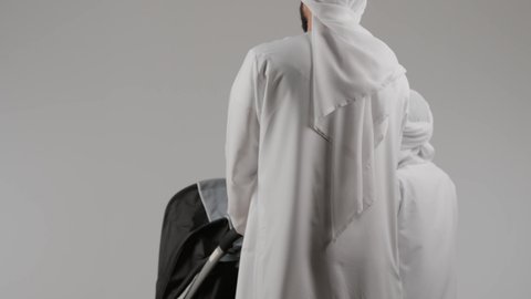 المشي والتنزه برفقة الأسرة، قضاء أوقات عائلية ممتعة ، بورتريه من الخلف  لرجل عربي خليجي اماراتي يرتدي الكندورة و الغتره يجر عربة الاطفال برفقة ابنه بإيماءات تدل على السعادة و المحبة