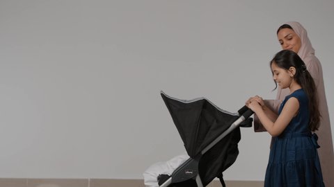 Spending fun family time, a family outing and tour, walking and hiking with the family, a portrait of an Emirati Gulf Arab woman wearing a colorful abaya, pulling a baby stroller with her little girl, with gestures indicating happiness, love and tenderness