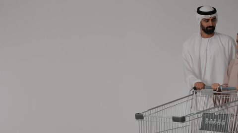 المشاركة في شراء المستلزمات المنزلية، الاستفادة من الخصومات والعروض، سيدة تشير بإصبع السبابة إلى شيء ما، بورتريه زوجان عربيان خليجيان اماراتيان يرتديان الزي التقليدي يقومان بجرعربة التسوق، شراء المنتجات من السوبر ماركت، التجول في المول والمركز التجاري