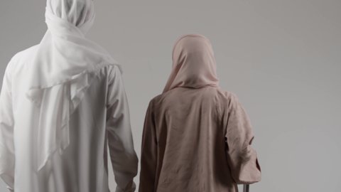 الذهاب في رحلة ممتعة، القيام بالجولات الترفيهية، السياحة في إجازة الصيف، بورترية من الخلف لزوجان عربيان خليجيان اماراتيان يرتديان الزي التقليدي يقومان  بالاستعداد لقضاء رحلة صيفية ترفيهية ممتعة، امراة خليجيه تمسك بيديها حقيبه السفر