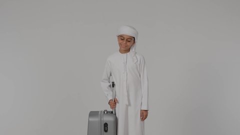 القيام بالجولات الترفيهية، مفهوم السياحة في إجازة الصيف،, طفلان عربيان خليجيان اماراتيان يقومان بالاستعداد  لقضاء رحلة صيفية ممتعة، طفل عربيى خليجي اماراتي يرتدي الكندورة و الغتره يمسك بيده حقيبه السفر بايماءات السعادة و السرور