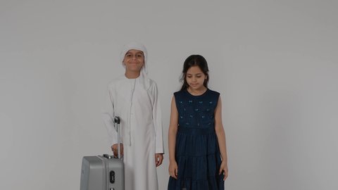 القيام بالجولات الترفيهية، مفهوم السياحة في إجازة الصيف، طفلان عربيان خليجيان اماراتيان يقومان بالاستعداد لقضاء رحلة صيفية ممتعة، طفل عربيى خليجي اماراتي يرتدي الكندورة و الغتره يمسك بيده حقيبه السفر بايماءات السعادة و السرور