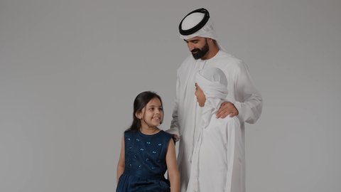 السياحة في إجازة الصيف, الشعور بالسعادة والفرح, عائله عربيه خليجيه اماراتية تستعد لقضاء رحلة صيفية ممتعة, بورترية لرجل عربي خليجي اماراتي يرتدي الكندورة و الغتره يقضي وقت ممتع مع اولادة ، طفله خليجيه تجلس على حقيبه السفر، خلفية رمادية