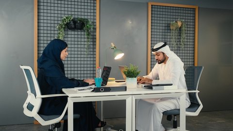 إنجاز الأعمال بسرعة وسهولة، أداء المهام والاعمال المكتبيه داخل مقر الشركه، استخدام التقنيات الحديثة المتطورة، تصوير من الجانب لشاب عربي خليجي اماراتي يرتدي الكندورة والغترة البيضاء يجلس فى المكتب و يقوم بالعمل على الحاسب المحمول برفقه زميلته