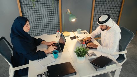 تحقيق أهداف الشركة، دراسه المشاريع ومراجعه التقارير السنويه، أداء المهام والاعمال المكتبيه داخل مقر الشركه، استخدام التقنيات الحديثة المتطورة، شاب عربي خليجي اماراتي يرتدي الكندورة والغترة البيضاء يجلس فى المكتب و يقوم بالعمل على الحاسب المحمول برفقه زميلته