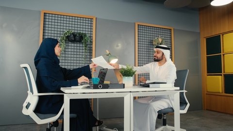 To cooperate and participate in completing work, studying projects and reviewing annual reports, performing tasks and office work within the company’s headquarters, using modern advanced technologies, a young Emirati Gulf Arab man wearing a white kandura and ghutra sitting in the office and working on the laptop with his colleague