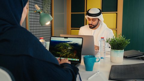 دراسه المشاريع ومراجعه التقارير السنويه، أداء المهام والاعمال المكتبيه داخل مقر الشركه، استخدام التقنيات الحديثة المتطورة، إدارة شؤون الأعمال وتنظيمها، شاب عربي خليجي اماراتي يرتدي الكندورة والغترة البيضاء يجلس فى المكتب و يقوم بالعمل على الحاسب المحمول برفقه زميلته