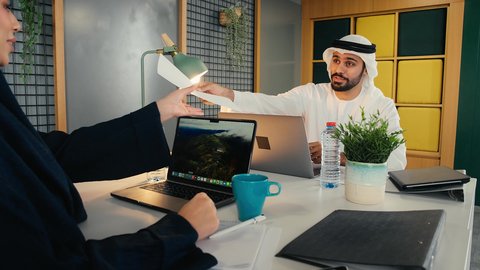 أداء المهام والاعمال المكتبيه داخل مقر الشركه، استخدام التقنيات الحديثة المتطورة، إدارة شؤون الأعمال وتنظيمها، شاب عربي خليجي اماراتي يرتدي الكندورة والغترة البيضاء يجلس فى المكتب و يقوم بالعمل على الحاسب المحمول برفقه زميلته, دراسه المشاريع ومراجعه التقارير السنويه