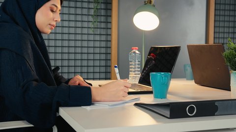 تحليل نتائج البيانات الاحصائية، تصوير مقرب لامراة عربية خليجية اماراتية محجبة ترتدي العباءة تجلس خلف الطاولة تقوم باستخدام الالة الحاسبة لاجراء العمليات الحسابية، انجاز المهام في المكتب، سيدة سعودية تدون الملاحظات على الورق، وظيفة مكتبية في شركات الخليج