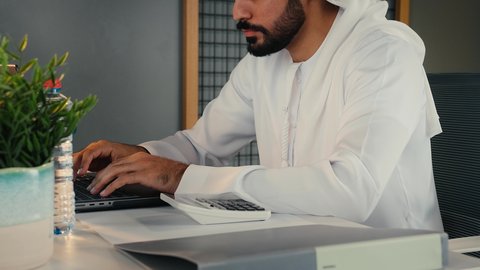 استخدام الالة الحاسبة لاجراء العمليات الحسابية, استخدام التقنيات الحديثة و المتطورة في العمل, مهن و وظائف في الامارات , تصوير مقرب من الجانب لشاب عربي خليجي اماراتي يرتدي الثوب التقليدي يجلس داخل المكتب يعمل على الحاسب المحمول بايماءات الاندماج و التركيز, أعمال إدارية مكتبية