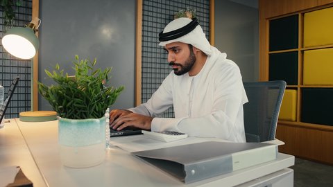 التواصل مع العملاء عن بعد, استخدام التقنيات الحديثة و المتطورة في العمل, مهن و وظائف في الامارات , تصويرمن الجانب لشاب عربي خليجي اماراتي يرتدي الثوب التقليدي يجلس داخل المكتب يعمل على الحاسب المحمول بايماءات الاندماج و التركيز, أعمال إدارية مكتبية