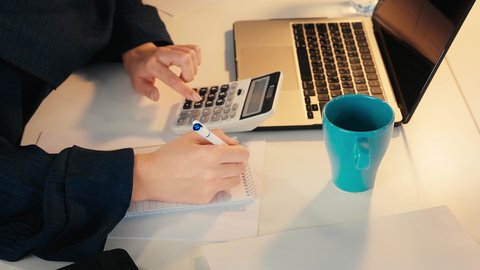 تحليل نتائج البيانات الاحصائية، تصوير مقرب ليد  امراة عربية خليجية اماراتية محجبة ترتدي العباءة تجلس خلف الطاولة تقوم باستخدام الالة الحاسبة لاجراء العمليات الحسابية، انجاز المهام في المكتب، سيدة سعودية تدون الملاحظات على الورق، وظيفة مكتبية في شركات الخليج