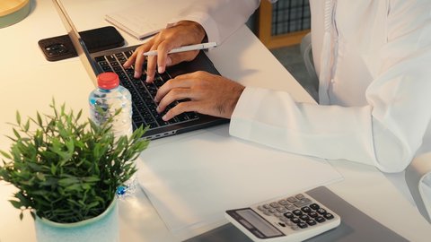 Taking notes on paper, office job in UAE companies, analyzing statistical data results, close-up of hand of an Emirati Gulf Arab man wearing kandura and ghutra sitting behind office table using laptop