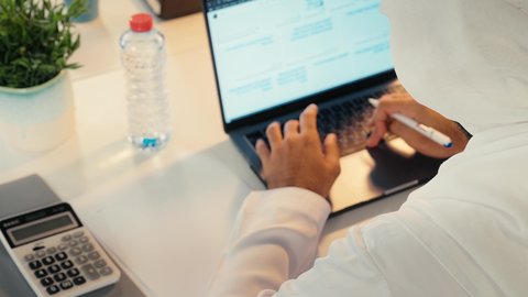 وظيفة مكتبية في شركات الامارات العربية المتحدة، تحليل نتائج البيانات الاحصائية، تصوير مقرب ليد رجل عربي خليجي اماراتي يرتدي الكندورة والغترة يجلس خلف طاولة المكتب يقوم باستخدام الحاسب المحمول ، تدوين الملاحظات على الورق