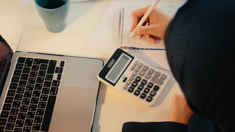 سيدة سعودية تدون الملاحظات على الورق، انجاز المهام في المكتب، وظيفة مكتبية في شركات الخليج، تحليل نتائج البيانات الاحصائية،تصوير مقرب لامراة عربية خليجية اماراتية محجبة ترتدي العباءة تجلس خلف الطاولة تقوم باستخدام الالة الحاسبة لاجراء العمليات الحسابية