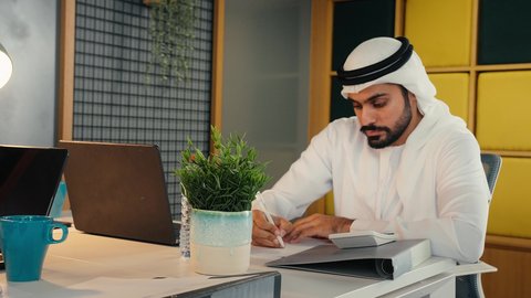 تحليل نتائج البيانات الاحصائية، رجل عربي خليجي اماراتي يرتدي الكندورة والغترة يجلس خلف طاولة المكتب ويعمل على الحاسب المحمول لانجاز المهام، تدوين الملاحظات على الورق، استخدام الالة الحاسبة لاجراء العمليات الحسابية، وظيفة مكتبية في شركات الامارات العربية المتحدة