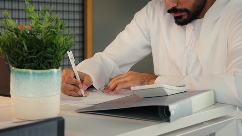 استخدام الالة الحاسبة لاجراء العمليات الحسابية، وظيفة مكتبية في شركات الامارات، تحليل نتائج البيانات الاحصائية، تدوين الملاحظات على الورق، احتساء القهوة في المكتب، تصوير مقرب لرجل عربي خليجي اماراتي يرتدي الكندورة يجلس في مكتبه ويقوم بإنجاز مهام العمل
