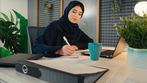 وظيفة مكتبية في شركات الخليج، انجاز المهام في المكتب، تحليل نتائج البيانات الاحصائية، تصوير لامراة عربية خليجية اماراتية محجبة ترتدي العباءة تجلس خلف الطاولة تقوم بكتابة التقارير، سيدة سعودية تدون الملاحظات على الورق