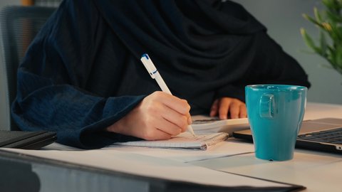 تحليل نتائج البيانات الاحصائية، تصوير مقرب  ليد امراة عربية خليجية اماراتية محجبة ترتدي العباءة تجلس خلف الطاولة تقوم بكتابة التقارير، انجاز المهام في المكتب، سيدة سعودية تدون الملاحظات على الورق، وظيفة مكتبية في شركات الخليج