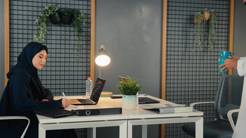 استخدام التقنيات الحديثة والتكنولوجيا بالمجال الإداري، التوظيف في المكاتب والشركات الاماراتية، امراة عربيه خليجيه اماراتيه ترتدي العباءة السوداء تجلس على المكتب وأمامها الحاسوب المحمول، رجل خليجي يقوم بتقديم كوب من المشروب الساخن لزميلته
