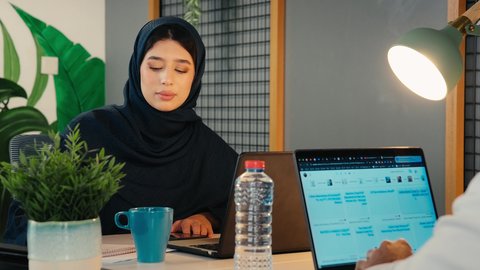 Employment in UAE offices and companies, use of modern techniques and technology in the administrative field, answering and responding to work phone calls via mobile, close-up of an Emirati Gulf Arab woman wearing a black abaya sitting at a desk with a laptop in front of her, enjoying a drink