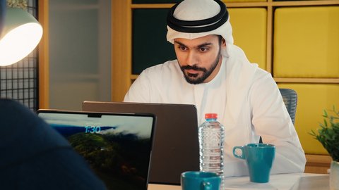 استخدام التقنيات الحديثة والتكنولوجيا بالمجال الإداري، التوظيف في المكاتب والشركات الاماراتية، الإجابة والرد على مكالمات العمل الهاتفية عبر الجوال، تصوير مقر لرجل عربي خليجي اماراتي يرتدي الثوب التقليدي والغترة يجلس على المكتب وأمامه الحاسوب المحمول