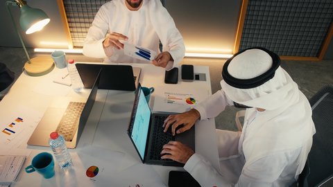 شرح و تحليل الرسوم البيانية، دراسه وتحليل المشاريع الجديده ومناقشه اداء وتقارير العمل، إدارة مهام العمل اليومية في مقر الشركة، تطوير الخطط والاسترتيجيات، تصوير مقرب لرجلان عربيان خليجيان اماراتيان يرتديان الكندورة و الغتره يجلسان في المكتب يقومان بالعمل على جهاز الحاسوب المحمول