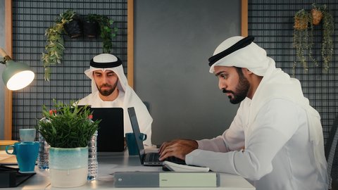 Studying and analyzing new projects, discussing performance and work reports, Emirati professions and jobs, managing daily work tasks at the company headquarters, developing plans and strategies, using modern technical devices, two Emirati Gulf Arab men wearing kandura and ghutra sitting in the office working on the laptop