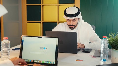 دراسة وتحليل المشاريع الجديده وتقارير العمل، مهن ووظائف مكتبية ، تصوير مقرب لرجل عربي خليجي اماراتي يرتدي الكندوره والغتره البيضاء الاماراتيه يجلس على المكتب بالعمل على جهاز الاب توب المحمول بايماءات الاندماج و التركيز
