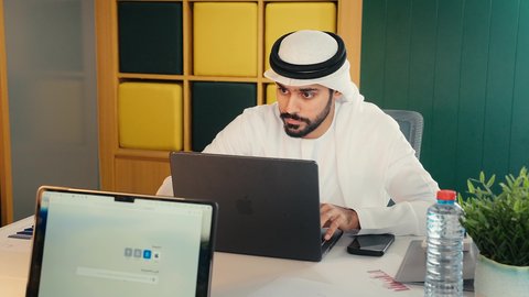 مهن ووظائف مكتبية ، تصوير مقرب لرجل عربي خليجي اماراتي يرتدي الكندوره والغتره البيضاء الاماراتيه يجلس على المكتب بالعمل على جهاز الاب توب المحمول بايماءات الاندماج و التركيز، دراسة وتحليل المشاريع الجديده  وتقارير العمل