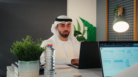 Completing work tasks, computer and information technology professionalism, close-up of an Emirati Gulf Arab man wearing traditional dress sitting in a work office, using a laptop, surfing the internet, office work, workplace, using modern technology