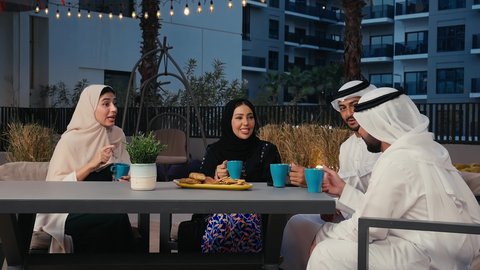 قضاء أوقات ممتعة، نقاشات وحوارات، صداقات وتعارف، الجلوس في الهواء الطلق، رجلان عربيان خليجيان اماراتيان يرتديان الزي الاماراتي التقليدي و امراتان عربيتان اماراتيان يستمتعون بتبادل الحديث في احد المقاهي الاماراتية، الاستمتاع بتناول المشروبات