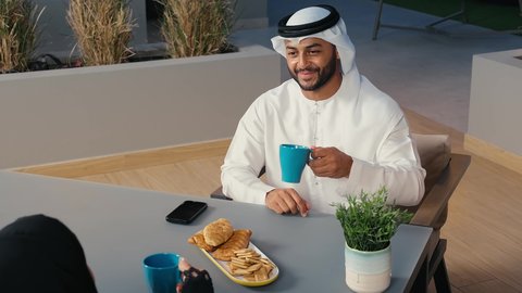 Enjoying cold drinks and delicious seasonal juices, spending enjoyable times outdoors in a pleasant atmosphere, going out on a family evening, an Emirati Gulf Arab man wearing a kandura and ghutra sitting in a cafe with his wife in the United Arab Emirates, strengthening marital relations and bonds of love and affection