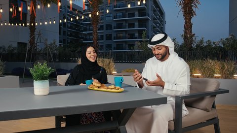 الاستمتاع بتناول المشروبات ، قضاء الأوقات الممتعة، توثيق اللحظات العائلية السعيدة, استخدام الأجهزة التقنية الحديثة, رجل عربي خليجي إماراتي يرتدي الكندورة و الغترة البيضاء يجلس فى مقهي يقوم بالتقاط صورة سيلفي عبر الهاتف المحمول برفقه زوجته ، ايماءات السعاده و السرور