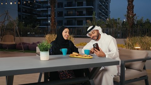 قضاء الأوقات الممتعة  توثيق اللحظات العائلية السعيدة, استخدام الأجهزة التقنية الحديثة, رجل عربي خليجي إماراتي يرتدي الكندورة و الغترة البيضاء يجلس فى مقهي و يشاهد الهاتف المحمول  برفقه زوحته, مطاعم و كافيهات في الإمارات