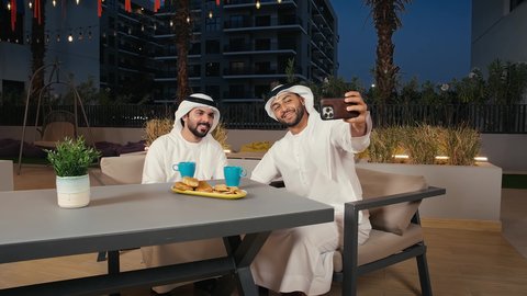 الاحتفاظ باللحظات السعيدة، تطوير العلاقات الإجتماعية، الاستمتاع بقضاء الوقت مع الأصدقاء، مفهوم التسلية والترفيه, تصوير لرجلان عربيان خليجيان اماراتيين يرتديان الكندورة و الغتره يقومان بالتقاط صور سيلفي تذكارية عبر الجوال فى احدي المقاهي بايماءات السعاده و السرور