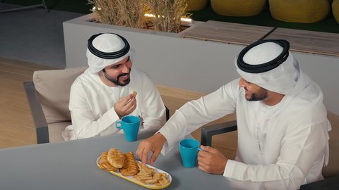 Having a quick meal and a refreshing drink, the concept of entertainment and leisure, spending a good time with friends, a close-up of two Emirati Gulf Arab men wearing kandura and ghutra exchanging conversations and discussions inside a cafe with gestures of pleasure and happiness